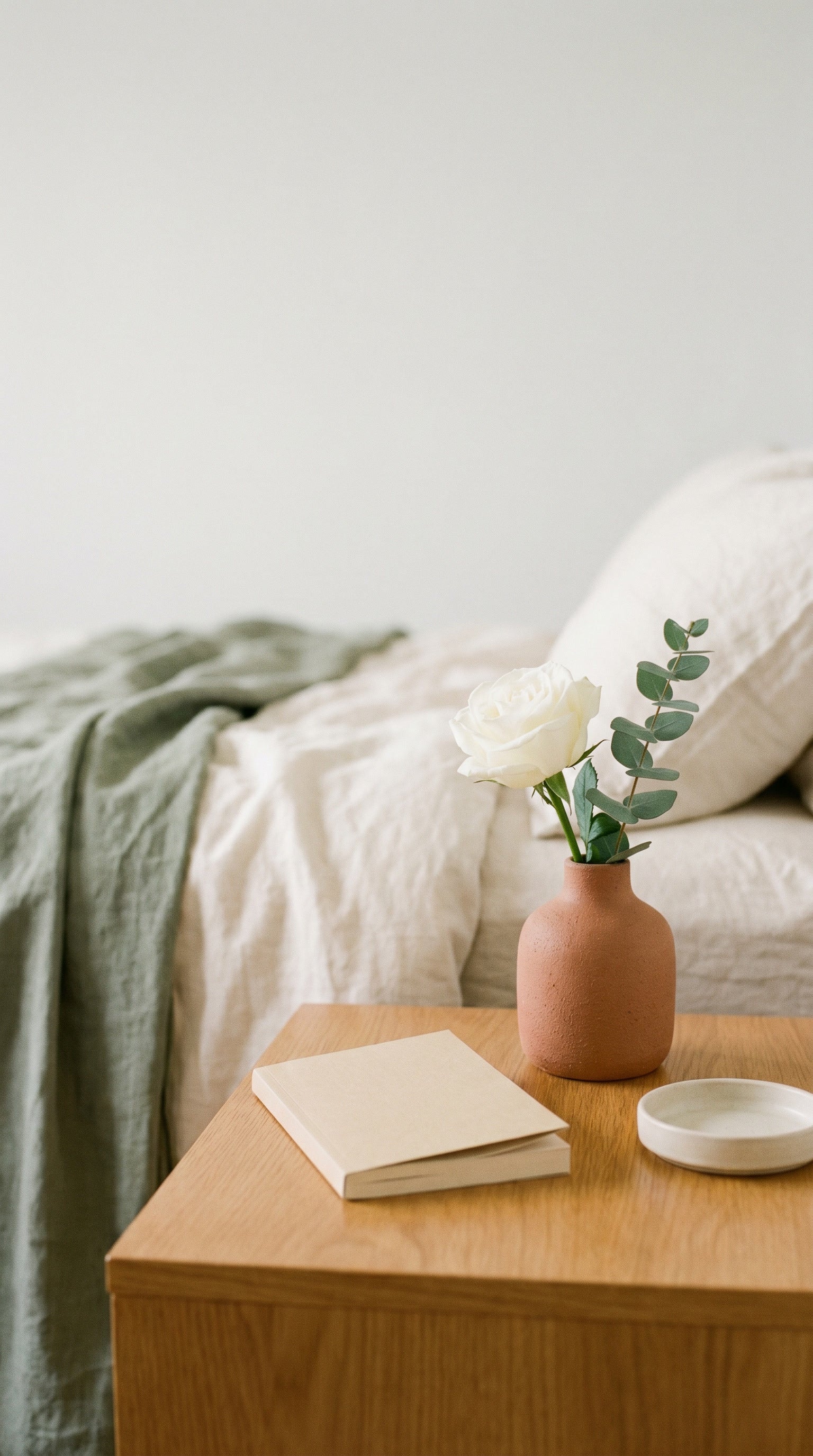 A single white rose with eucalyptus in a small terracotta vase on a bedside table, beside a notebook and a saucer